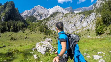 A man taking a selfie while hiking in lush green Alpine region in Austria. The sky is blue, with one cloud on it. Mountains slopes overgrown with bushes and grass. Summer in Alps. Exploring the nature