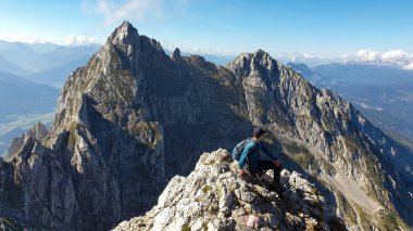 A man sitting at the edge of a mountain with a view on a vast valley. There are sharp mountains and high peaks around. The Alpine slopes are almost barren. Lush green valley. Bright day. Freedom.