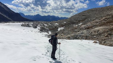 A man with hiking stick hiking through a glacier in Heiligenblut region in Austria, on the way to Hohe Sonnblick. There are many high chains of Alps behind him. Sunny day. Adventure and discovery.
