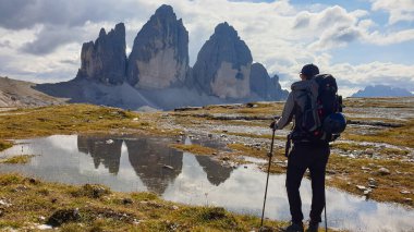 A man in hiking outfit enjoying the view on the famous Tre Cime di Lavaredo (Drei Zinnen), mountains in Italian Dolomites. The mountains are reflecting in small paddle. Desolated and raw landscape.