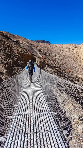 A man crossing through a suspension bridge in Himalayas. The valley is located in Mustang region, Annapurna Circuit Trek in Nepal. The man carried a heavy backpack. Barren slopes. Harsh landscape.