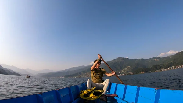 A man in yellow safety jacket, sitting in a blue boat and paddling across Phewa Lake in Pokhara, Nepal. Behind him there are high, snow capped Himalayas with Mt Fishtail (Machhapuchhare) between them