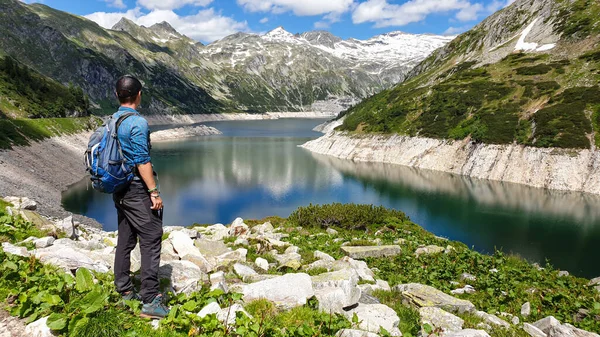 A man with hiking backpack standing at the side of an artificial lake at a dam in Austria. The lake has navy blue color. High Alps around. There is a glacier in the back. Adventure and discovering