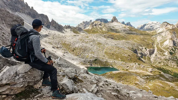 A man with hiking backpack and sticks enjoying the view on Italian Dolomites. There are sharp and steep mountains. At the bottom of a small valley there is a small, navy blue lake. Raw landscape