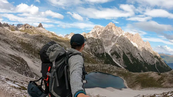 A man with hiking backpack and sticks taking a selfie in Italian Dolomites. There are sharp and steep mountains. At the bottom of a small valley there is a small, navy blue lake. Raw landscape. Remedy