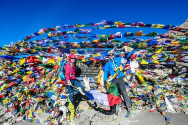 A couple standing between prayer flags at the top of Thorung La Pass, Annapurna Circuit Trek, Nepal. Memorial board. Colorful prayer flags attached to the stone wall, blown by the wind. Clear sky.
