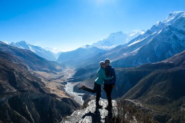 A couple standing at a mountain ledge, hugging and enjoying the view on Manang valley stretching in Himalayas, along Annapurna Circuit. Freedom. Love and passion. Snow capped Himalayas around.