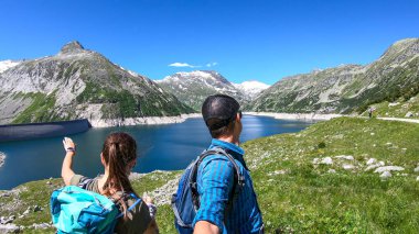 A couple with hiking backpacks standing at the side of an artificial lake at a dam in Austria. The lake has navy blue color. High Alps around. There is a glacier in the back. Adventure and discovering