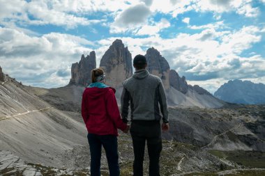 A couple in hiking outfit holding hands and enjoying the view on the famous Tre Cime di Lavaredo (Drei Zinnen) in Italian Dolomites. Desolated and raw landscape, full of lose stones. Overcast. Love