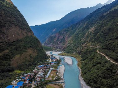 A view from above on the Tal Village in Himalayas, Nepal.  High mountains guarding the village, crystal clear river going through the village. Sun is slowly setting. Lots of blue rooftops.