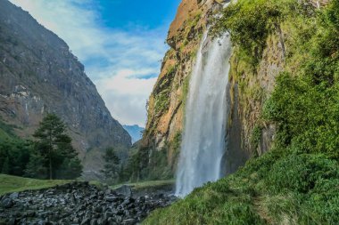 A waterfall spotted in Tal, Annapurna Circuit Trek, Nepal. Few hundred meters of free fall, waterfall surrounded by tall mountains slopes, covered with green bushes and trees. Smooth capture