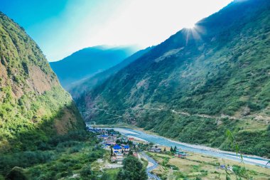 Tal village seen from above, Annapurna Circuit Trek, Nepal. Sun sets behind the high mountain, leaving the valley in the shadow. turquoise river flows next to the houses with blue roofs. 