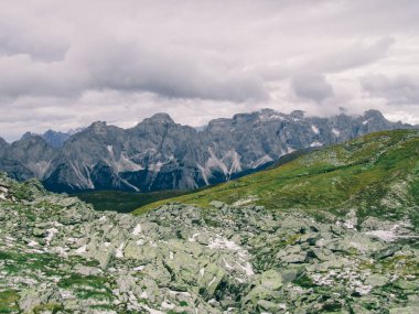 Alpine landscape of Carnic Alps. Lush green meadows. Stony hills, with leftovers of snow. In the background steep, sharp peaks. Huge overcast. No human interference in the area. Natural habitat