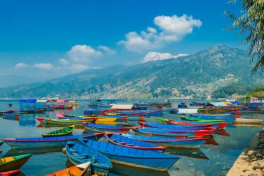 Many colourful boats parked on the shore of Phewa lake, Pokhara, Nepal. In the back Himalayan mountain range. Small villages visible on the shore. Peaceful and chilled atmosphere. Place to relax.