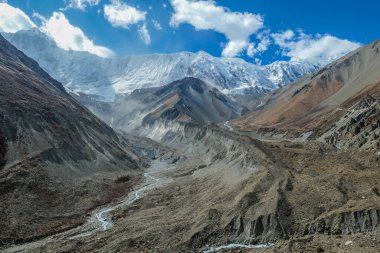 Himalayas, Annapurna Circuit Trek, Himalayas, Nepal. Dry and barren slopes. Steep hiking trail. Dangerous landscape. High and powerful mountains in the back. Overcast, with parts of blue sky. 