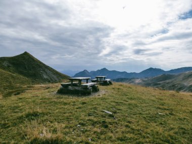 A resting place on the meadow in Millstatt, Austria. Two tables and 4 benches surrounded by grassland and high mountain peaks. Clouds. Calmness and emptiness. Hikers resting area.