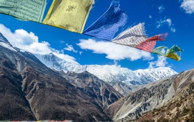 Manang/Nepal - 10 30 2018: Waving flags with mantra 'Om mani padme hum' on them. Wind blows them over Himalayan peaks. Very weary flags. High peaks covered with snow. meditation and retreat