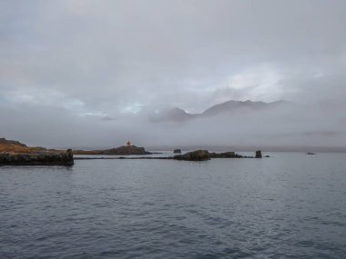 A shore of a fjord. At one of the headlands there is a small orange lighthouse. In the back higher mountains hiding in the clouds. Calm water surrounding the headlands.