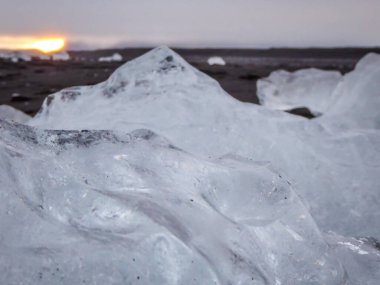 A close up on a piece of an ice berg. The surface of the ice is crystal-transparent. Ice berg placed on a black sand beach. In the back sun sets down. Romantic ans serene atmosphere. Cold temperature