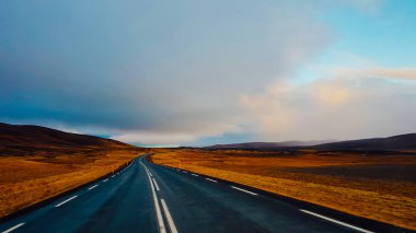 An endless road through the lowlands. On the side some hills. Dark and heavy tones. Both sides of the road are barren. Sky is clouded, but still blue parts visible. Empty road, not a single car moving