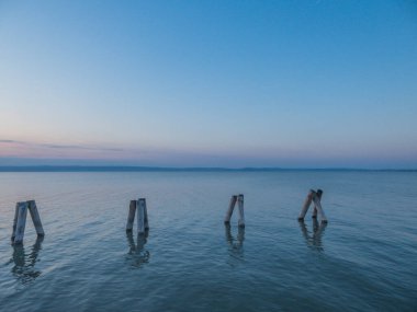 Soft colors of a sunset on the lake. pillars coming out of the calm water. Pillars in groups of three, form shapes of  pyramids. Calm surface of the lake. Mountains in the back. Pinky sky.