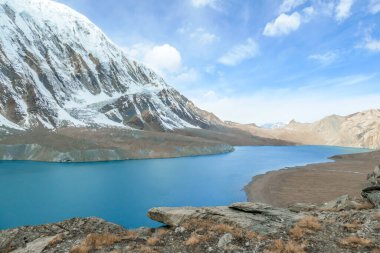 Tilicho Lake, Annapurna Circuit Trek detour, Nepal. Blue and calm surface of the lake, mountains covered in the shadow, sunlight in the back. Overcast. Harsh landscape. Thankfulness, freedom.