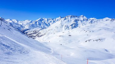 A beautiful and serene landscape of mountains covered with snow. Thick snow covers the slopes. Clear weather. Perfectly groomed slopes. Massive ski resort. Red ski poles on the side of the trail.