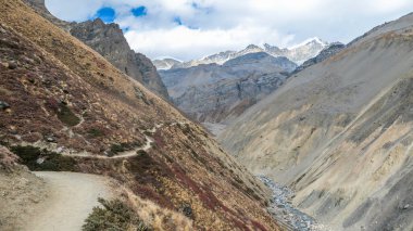 endless trekking trail among sharp peaks. Blue sky with overcast. Early morning, waking up to natural wonder. Annapurna Circuit Trek, Nepal. Dry grass. Other side of the gorge very raw and barren.