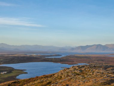 A flatland, covered with golden grass. Dried grass due to the winter season. Fjord view, with some taller mountains in the back. Calm water of the fjord. Clear weather with some clouds on the sky.