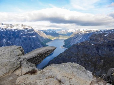 Famous rock formation, Trolltunga with a view from the above on Ringedalsvatnet lake, Norway. Rock hanging. Slopes of the mountains are partially covered with snow. The water of the lake is navy blue.