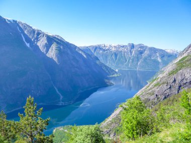 An majestic view on Eidfjord from Kjeasen, Norway. Slopes of the mountains are overgrown with lush green grass. Water has dark blue color. Taller parts of the mountains are barren. Sunny and clear day