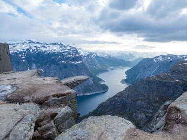 Famous rock formation, Trolltunga with a view from the above on Ringedalsvatnet lake, Norway. Rock hanging. Slopes of the mountains are partially covered with snow. The water of the lake is navy blue.