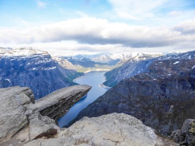 Famous rock formation, Trolltunga with a view from the above on Ringedalsvatnet lake, Norway. Rock hanging. Slopes of the mountains are partially covered with snow. The water of the lake is navy blue.