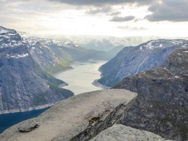 Famous rock formation, Trolltunga with a view from the above on Ringedalsvatnet lake, Norway. Rock hanging. Slopes of the mountains are partially covered with snow. The water of the lake is navy blue.