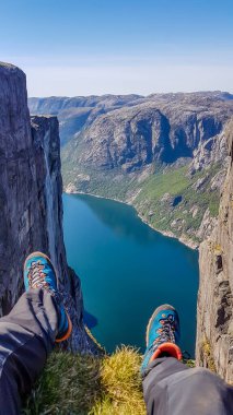 Two feet with hiking boots on them hanging down from a steep rock with a view on stunning Lysefjorden shimmering with many shades of blue and green. Steep slopes of the mountain,waterfall rushing down