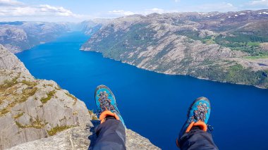 Two feet with hiking boots on them hanging down from a steep rock with a view on stunning Lysefjorden shimmering with many shades of blue and green. Steep slopes of the mountain,waterfall rushing down