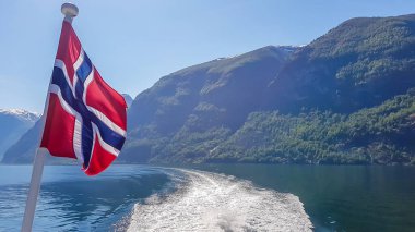 Norwegian flag hanging on  the railing of the ship and waving above the water.The motor of the ship makes the water wavy and foamy. Tall, lush green mountains surrounding the fjord. Clear blue sky.