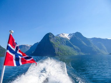 Norwegian flag hanging on  the railing of the ship and waving above the water.The motor of the ship makes the water wavy and foamy. Tall, lush green mountains surrounding the fjord. Clear blue sky.