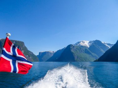 Norwegian flag hanging on  the railing of the ship and waving above the water.The motor of the ship makes the water wavy and foamy. Tall, lush green mountains surrounding the fjord. Clear blue sky.