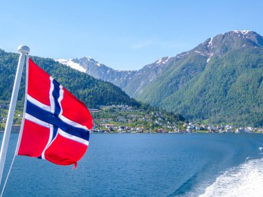Norwegian flag hanging on  the railing of the ship and waving above the water.The motor of the ship makes the water wavy and foamy. Tall, lush green mountains surrounding the fjord. Clear blue sky.
