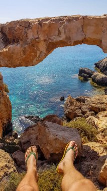 A pair of flip flops ahanging down from a steep cliff, with a stony arch in the back. Cape Greco, Cyprus, bridge connecting two cliffs. Barren slopes of the stony formation Endless line of a horizon