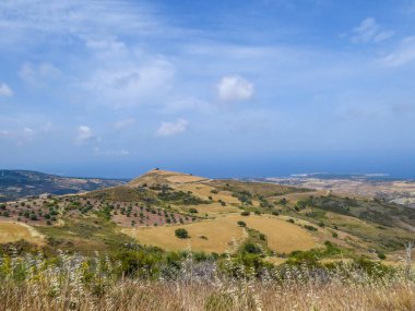 An idyllic view of a hilly fields in Cyprus. Slopes are overgrown with grains and small bushes. In the back some smaller mountains are visible. Great overcast, spreading all over the sky.
