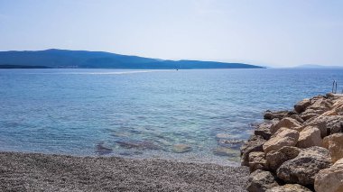 Stony beach in Krk, Croatia. The sea has a turquoise color. Surface of the sea is calm, there are no waves. In the back there are some mountains. A pile of bigger stones on the side. A bit of overcast