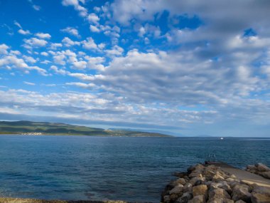 Calm sea slowly moving towards the shore. The surface of the water is almost still. In the back there are some islands overgrown with small bushes. In front a big rock emerging from the sea