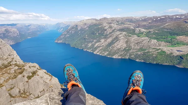 Two feet with hiking boots on them hanging down from a steep cliff with a view on stunning Lysefjorden shimmering with many shades of blue and green. View from Preikestolen. Clear and bright day.