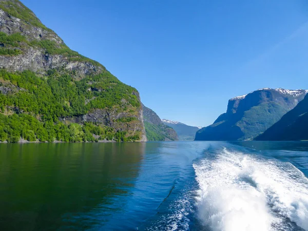 A view on the Songefjorden fjord from the water level. The motor of the ship makes the water wavy and foamy. Tall, lush green mountains surrounding the fjord. Clear blue sky.