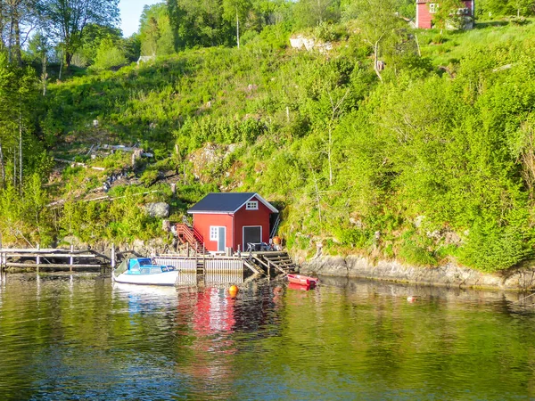 A little red house built by the shore of a Songefjorden, Norway, seen from the boat. Tall, lush green mountains surrounding the little house. Calm surface of the water.