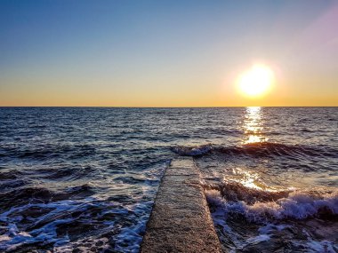 Romantic sunset by a stony seashore. The sun sets over the horizon. The sun beams reflecting in the calm sea waters. Stony shore is washed by the gentle waves. The sky is turning yellow and orange.