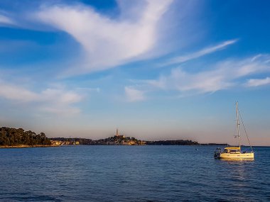 Golden hour in the harbor next to Rovinj, Croatia. The city is located in the back. A small yacht anchored to a buoy. Soft clouds on the sky. Sea water is very calm.