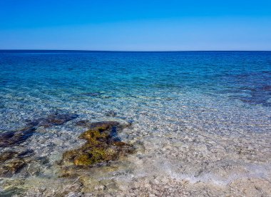 Calm, shallow sea water. There are barely any waves on the surface. The water is very clear, one can see the stony bottom. Few bigger stones protrude from water.